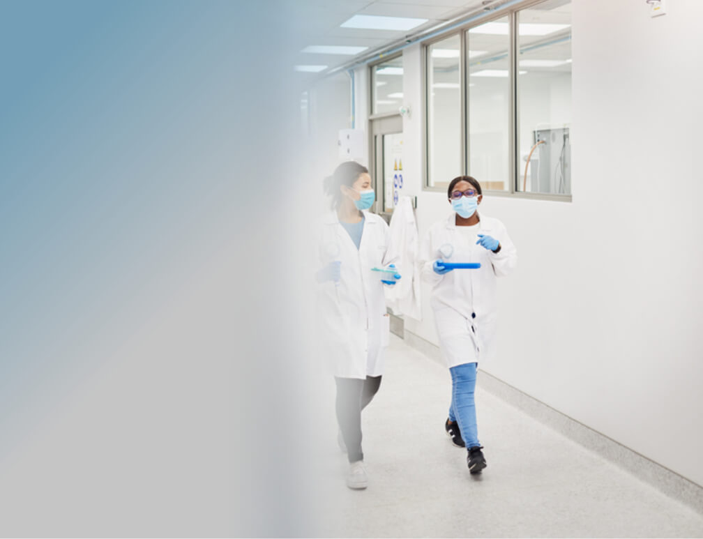 Two masked scientists in protective attire walk down a brightly lit laboratory corridor.