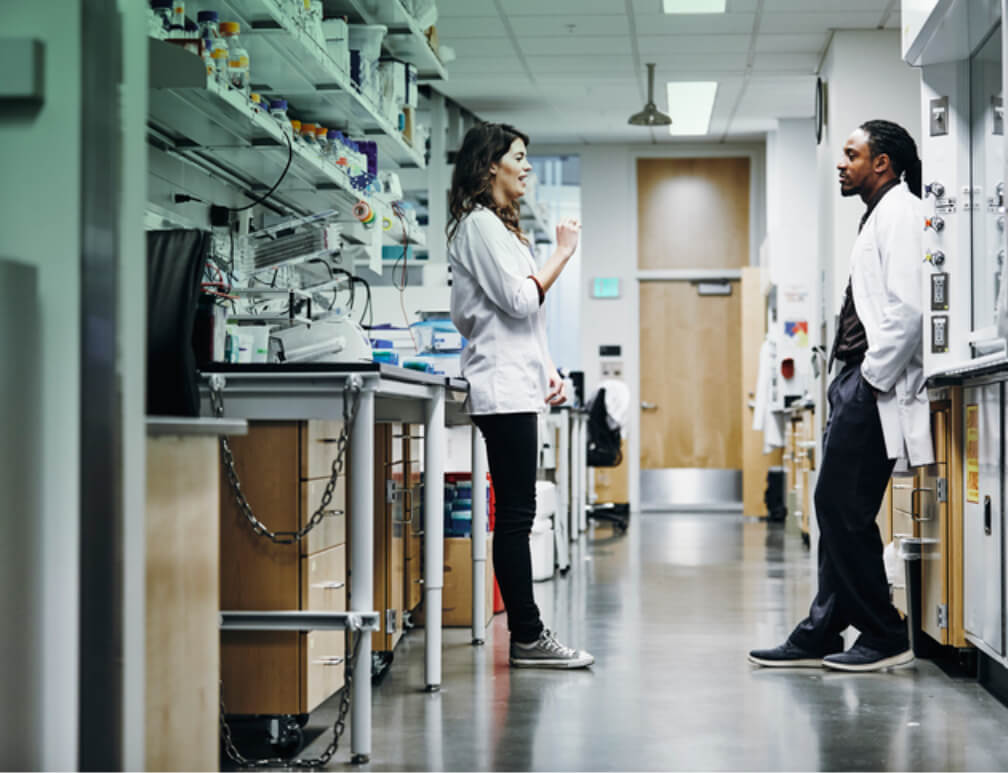 Two scientists in lab coats discuss research in laboratory.